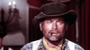 A rugged man in a cowboy hat, plaid shirt, and yellow scarf with a cigar stares ahead; the dimly lit background and red curtains evoke a classic western mood—just like on "The Last Gun (1964) DVD.
