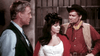 Three people in Western attire stand indoors: a man in a dark vest faces a woman in a white dress and red shawl, with another man in cowboy hat and red shirt beside her. Jail bars suggest a classic scene from The Last Gun (1964) DVD.