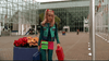 A young woman with blonde pigtails strolls outside a glass building, carrying bright red and blue suitcases that evoke the vibrant style of 90s nostalgia, much like Zenon Girl of the 21st Century (1999) DVD. She sports a lively ensemble in green, blue, and pink hues—an outfit that looks as if it came straight from the world of Zenon herself. Nearby, large planters overflow with red and yellow flowers.