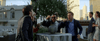 Four men excitedly gather around a table outdoors, enthusiastically eating from bowls while discussing their favorite scenes from “A California Christmas: City Lights (2021)” on DVD. In the background, three people stand talking and holding drinks, with city buildings visible.