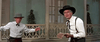 Two men in cowboy hats and western attire stand outside a building with a wooden railing. One, holding a book and looking surprised, faces the other who gestures animatedly—evoking the adventure of Ride A Crooked Trail (1958) DVD.