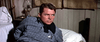 A man in a gray plaid shirt with a sheriff's badge reclines on a bed, evoking the Western adventure of Ride A Crooked Trail (1958) DVD. An oil lamp and brown curtains are visible behind him as he looks to his left, serious.