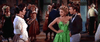 At a lively gathering, guests in colorful dresses and suits mingle as a woman in white faces a couple; the woman wears a bright green dress inspired by classic Western style, reminiscent of "Ride A Crooked Trail (1958)" DVD.