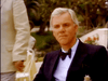 A gray-haired man in a black tuxedo and bow tie sits outdoors, looking serious, as if attending a gathering for Monte Carlo (1986) DVD fans. Nearby, someone in a light suit stands amid greenery and flowers.