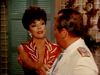 A woman in a red dress and a man in a white suit with navy accents stand amid green shutters and plants, evoking the glamour of classic TV miniseries "Monte Carlo (1986)," now available on DVD.