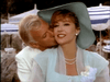A man in a white suit kisses a smiling woman in a light blue hat and dress, pearls around her neck. Outdoors with striped umbrellas, the scene recalls the elegance of classic TV as seen in the Monte Carlo (1986) DVD.