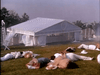 Several people in white lie motionless on grass near a white open tent, creating a dramatic scene reminiscent of the "Monte Carlo (1986) DVD," with scattered hats and a hazy background.