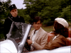 A police officer stands beside a convertible, talking to a man in a beige suit and a woman in a hat—just like scenes from the rare "Monte Carlo (1986) DVD"—with trees in the background.
