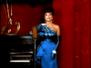 A woman in a glamorous blue, one-shoulder gown stands by a piano onstage with red curtains, evoking classic TV miniseries elegance—just like scenes from the "Monte Carlo (1986) DVD"—as she faces the audience, mic in hand.
