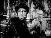 A man in a dark hat and glasses sits at a bar as the bartender pours a drink behind him. This black-and-white scene captures the vintage charm of "The Slowest Gun in the West (1960)" DVD.