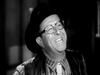 A man in glasses, a dark hat, and a striped shirt smiles broadly in this black-and-white image, capturing the joyful spirit of "The Slowest Gun in the West (1960)" DVD—like a star from classic comedy.