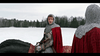 A knight in chainmail and a red cloak sits astride a horse in a snowy field, gazing at another similarly dressed figure nearby; with trees bordering the scene, the atmosphere captures the magical spirit of “The Knight Before Christmas (2019) DVD.”.
