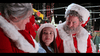 A young girl in a white knit hat sits between two adults dressed as Santa and Mrs. Claus, all smiling warmly amid holiday lights and festive decorations outdoors at night—creating a scene reminiscent of *The Knight Before Christmas (2019) DVD*.
