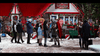 People gather outside a festive, snow-covered bakery adorned with Christmas lights and candy canes. A man dressed in shining knight armor converses with a woman in a gray coat, as though they’ve emerged straight from The Knight Before Christmas (2019) DVD. Surrounded by holiday decorations and gingerbread figures, the scene perfectly captures the enchanting spirit of a magical Netflix holiday romance.