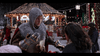 A man in medieval armor stands in front of a festive, illuminated gazebo at night, holding something and talking to a woman. Snow drifts gently down while people gather to enjoy the holiday decorations and sparkling lights—recreating a magical scene straight from “The Knight Before Christmas (2019)” DVD.