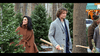 A woman in a brown coat and a man in a gray coat holding an axe walk among Christmas trees in a snowy forest, with other people in the background, evoking the enchanting atmosphere of “The Knight Before Christmas (2019)” DVD.