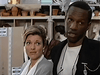 A woman and a man stand indoors, surrounded by shelves displaying pots, mannequin heads, and a selection of classic movies on DVD—including The Counterfeit Contessa (1994). The woman looks ahead with a slight smile, while the man—sporting a stethoscope around his neck—gazes thoughtfully to the side.