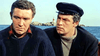 Two men sit in a boat on the water; one gazes downcast while the other, in a dark cap, looks his way. With a rocky island and towering lighthouse forming an imposing backdrop, this moment seems to leap straight from *Gorgo (1961)*—a rare classic featured on DVD and coveted by monster movie collectors.