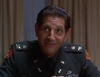 A man in a US military uniform with various badges and insignia sits at a table, smiling slightly, with beige curtains in the background—evoking the commanding presence and suspense found in "Breach of Conduct (1994)" on DVD.