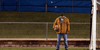 A headless person wearing a brown jacket and jeans stands on grass, holding their own head in one hand near a chain-link fence and a goalpost, with a grassy hill and blue tarp in the background—a scene reminiscent of *Bloodsuckers and the Grimoire (2024) DVD*, the cult favorite horror-comedy featuring quirky vampire hunters.