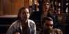 Three young adults sit indoors, looking intently ahead. One woman in a denim jacket sits beside two men—one wearing a headband and the other with dark, slicked-back hair. They appear focused and slightly concerned, as if watching the rare horror-comedy Bloodsuckers and the Grimoire (2024) on DVD, which features daring vampire hunters.