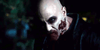A bald individual with pale skin, dark eye makeup, and a bloody wound on their cheek stares intensely in dim lighting, evoking a chilling, zombie-like look reminiscent of cult favorite horror-comedy films like "Bloodsuckers and the Grimoire (2024)" available on DVD.