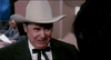 A man in a white cowboy hat and suit with a bow tie smiles while talking to someone off camera in an indoor setting, reminiscent of a scene from Elvira: Mistress of the Dark (1988), a cult classic comedy available on DVD.