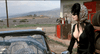 A woman in a low-cut black outfit and headscarf walks toward a vintage car with animal print seats, parked near an old gas station in a rural, hilly area—evoking the campy allure and cult-classic comedy of Elvira: Mistress of the Dark (1988) DVD.