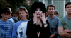 A woman with striking black hair and bold makeup—a clear nod to Elvira from “Elvira: Mistress of the Dark (1988)”—holds her hands in shock amid a group of surprised, paint-splattered people outdoors.