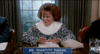 A woman with striking red hair and a crisp white collar sits at a table, smiling and gesturing. The nameplate in front of her reads “Ms. Chastity Pariah, Council Chairperson.” On the table are a glass of water and a beige purse—she looks every bit like someone you’d find debating rare DVDs, or even as if she stepped straight out of a cult classic comedy like *Elvira: Mistress of the Dark (1988)* on DVD.