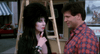 A woman with dramatic black hair and makeup—reminiscent of the cover art from the Elvira: Mistress of the Dark (1988) DVD—holds a finger to her lips, standing next to a man in a plaid shirt who is smiling at her. A ladder and a storefront can be seen in the background, channeling scenes from cult classic comedy films and rare movies available on DVD.