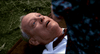 An older man with gray hair lies on the grass, looking up with a shocked, wide-eyed expression—like he’s just witnessed something outlandishly bizarre from *Elvira: Mistress of the Dark (1988)*. Part of another person in patterned clothing is visible in the foreground, reminiscent of a memorable scene from this cult classic comedy, now available on DVD.