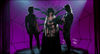 A woman dressed as Elvira: Mistress of the Dark stands center stage holding flowers, flanked by two men in minimal costumes, with a large spiderweb and purple lighting in the background—a striking scene reminiscent of the cult classic comedy, now available on DVD as “Elvira: Mistress of the Dark (1988).”.