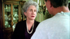An older woman with gray hair and a pearl necklace looks concerned while talking to a man in a white shirt in a dining room with a china cabinet in the background, as if they are quietly discussing the latest revelations covered in “Never Let Her Go (2001)” DVD—a story recently brought back into the spotlight by true crime miniseries.