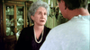 An older woman with gray hair and a pearl necklace looks concerned while speaking to a man in a softly lit room with a wooden cabinet and chandelier in the background, capturing the tense atmosphere depicted in *And Never Let Her Go (2001) DVD*.