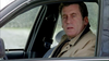 A man in a tan coat sits in the driver’s seat of a car, gazing thoughtfully out the open window with a serious expression, evoking the suspenseful mood of “Never Let Her Go (2001)” DVD. The car’s interior and a blurred outdoor background complete the cinematic scene.