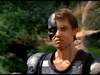 A man in armor stands outdoors, part of his face concealed by a metallic, robotic mask with a glowing red eye—evocative of the cyborg from the 1986 film "Eliminators" (DVD). The backdrop features green and rocky terrain.