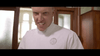 A person in a white medical coat with a stern expression stands indoors, looking directly at the camera. Wooden doors with frosted glass panels and sunlight in the background evoke the suspenseful atmosphere found in classic scenes from The Frighteners (1996) DVD.
