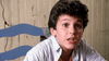 A young boy with curly dark hair and a striped shirt sits in front of a wooden wall, looking slightly upward with a surprised expression—like he’s just discovered a copy of the Little Monsters (1989) DVD on the shelf. A blue chair is visible behind him.