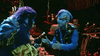 Two people in blue monster costumes with textured skin and horns talk animatedly in a dimly lit room filled with scattered food, candles, and shelves lined with rare DVDs—including “Little Monsters (1989)”—a true haven for any cult favorite film enthusiast.