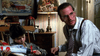 A boy sits holding a toy plane while an adult man with glasses and a tie leans toward him. They are indoors, with a decorated lamp and a painting in the background—creating an atmosphere reminiscent of scenes from the *Little Monsters (1989) DVD*.