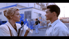 A woman holding folders talks to a man outside near a graffiti-covered wall, possibly discussing the “Only the Strong (1993) DVD.” Another person walks by in the background near a white building.