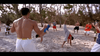 A group practices capoeira on a sandy beach, forming a circle reminiscent of scenes from “Only the Strong (1993)” DVD. Two individuals perform cartwheels in the center while others observe, with trees and sunlight in the background.