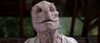 A humanoid creature with pink, scaly, lizard-like skin and small spikes on its head is wearing a white garment, standing indoors before blurred windows and greenery—resembling an eerie character you might discover in “Bad Fish (2024) DVD,” a maritime horror film prized by collectors.