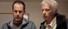 Two men stand side by side indoors, both looking down with serious expressions. One has dark hair and a beard, wearing a gray sweater; the other has white hair and wears a light shirt with a dark jacket. This scene captures the tense atmosphere of “Bad Fish (2024) DVD,” an ideal choice for fans of survival thriller DVDs seeking gripping narratives and unforgettable performances.