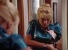 A woman with blonde hair in an updo stands by a mirror, adjusting her shiny blue floral dress and looking down with a serious expression—capturing a moment reminiscent of scenes from Kate’s Secret (1986) on DVD.