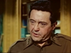 A man in a brown military uniform with epaulets is indoors, looking serious, as if captured in a suspense drama or classic World War II movie. Shelves and a window are blurred in the background, evoking the atmosphere of a scene from Fireball Forward DVD (1972).