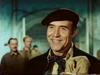 A smiling man wearing a dark beret, neckerchief, and coat stands indoors in a scene from the Fireball Forward DVD (1972), evoking the classic style of a suspense drama while several blurred people appear in the background.