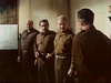 Four men in brown military uniforms stand indoors, engaged in a serious discussion in this suspenseful drama from Fireball Forward DVD (1972). Three of them face the man on the right, whose back is turned to the camera. A large map displayed on the left wall suggests strategic planning, evoking classic World War II scenes central to Fireball Forward.