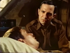 A man in military uniform sits beside another man who is lying in bed, appearing unwell. The man sitting looks concerned, and they are in a dimly lit room with brown tones—a tense atmosphere that evokes the classic suspense and wartime drama found in Fireball Forward DVD (1972).